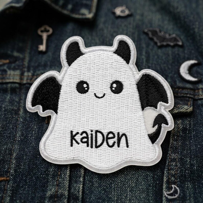 May include: An embroidered patch featuring a white ghost character with black bat wings, black ears, and a black tail. The ghost has a smiling face with two round black eyes and the name Kaiden stitched in black lettering across the centre.