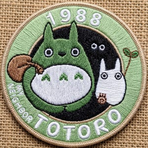 May include: A circular embroidered patch featuring a green Totoro character with a white belly, a smaller white Totoro, and black soot sprites. The text 1988 and MY NEIGHBOR TOTORO are stitched in white thread against a light green background.