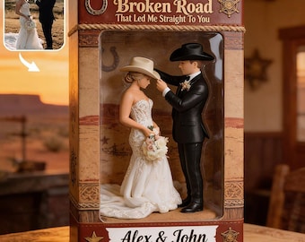 May include: A wedding cake topper figurine set inside a rectangular box featuring a bride in a white gown and a groom in a black suit and cowboy hat. The box displays the text God Bless The Broken Road That Led Me Straight To You and Alex & John.