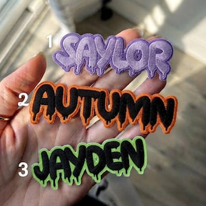 May include: Three embroidered name patches with a dripping font style. Patch 1 features the name SAYLOR in light purple. Patch 2 features the name AUTUMN in black with an orange border. Patch 3 features the name JAYDEN in black with a lime green border.