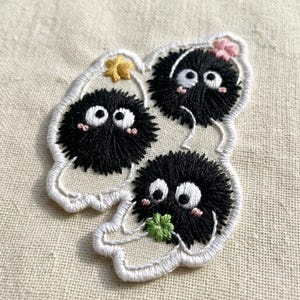 May include: An embroidered patch featuring three round, fuzzy black soot sprites with large white eyes and pink cheeks. One sprite holds a yellow star, one a pink flower, and one a green clover, all outlined in white thread on a fabric base.