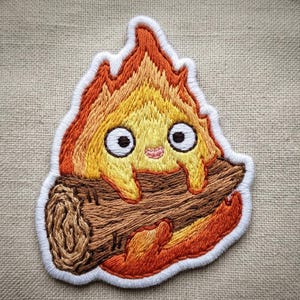 May include: An embroidered patch featuring a stylized flame character with large white eyes and a small pink mouth, holding a brown textured log. The flame is colored in shades of yellow and orange with a thick white border surrounding the entire shape.