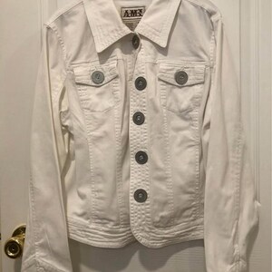 May include: A white denim jacket featuring a pointed collar, long sleeves with buttoned cuffs, and two chest pockets with button flaps. The front closure consists of a vertical row of large, round, metallic-toned buttons. The label reads AMX.