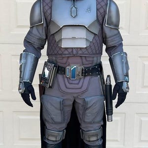 Op de afbeelding: Een volledig kostuum dat lijkt op een Mandalorian-krijger. De bepantsering is voornamelijk grijs en zilver, met een helm, borstplaat, schouderbeschermers en handschoenen. Het kostuum bevat een gereedschapsriem, zwarte handschoenen en laarzen. Een zwarte cape hangt aan de achterkant.