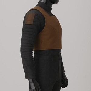 May include: A black long-sleeved shirt with a high neck and horizontal quilted detailing on the sleeves, worn under a cropped brown vest. The vest features a square neckline and a structured, solid design, paired with black trousers on a mannequin.