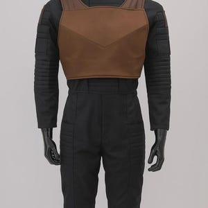 May include: A black long-sleeved jumpsuit featuring a high neck and ribbed detailing on the shoulders and thighs. Over the chest is a brown, structured, V-shaped vest piece with wide shoulder straps, worn on a glossy black mannequin torso.