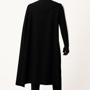 May include: A black long-line coat with a high collar and a cape-like back panel, worn over matching black trousers. The ensemble includes black gloves and black boots, displayed on a mannequin against a plain white background.