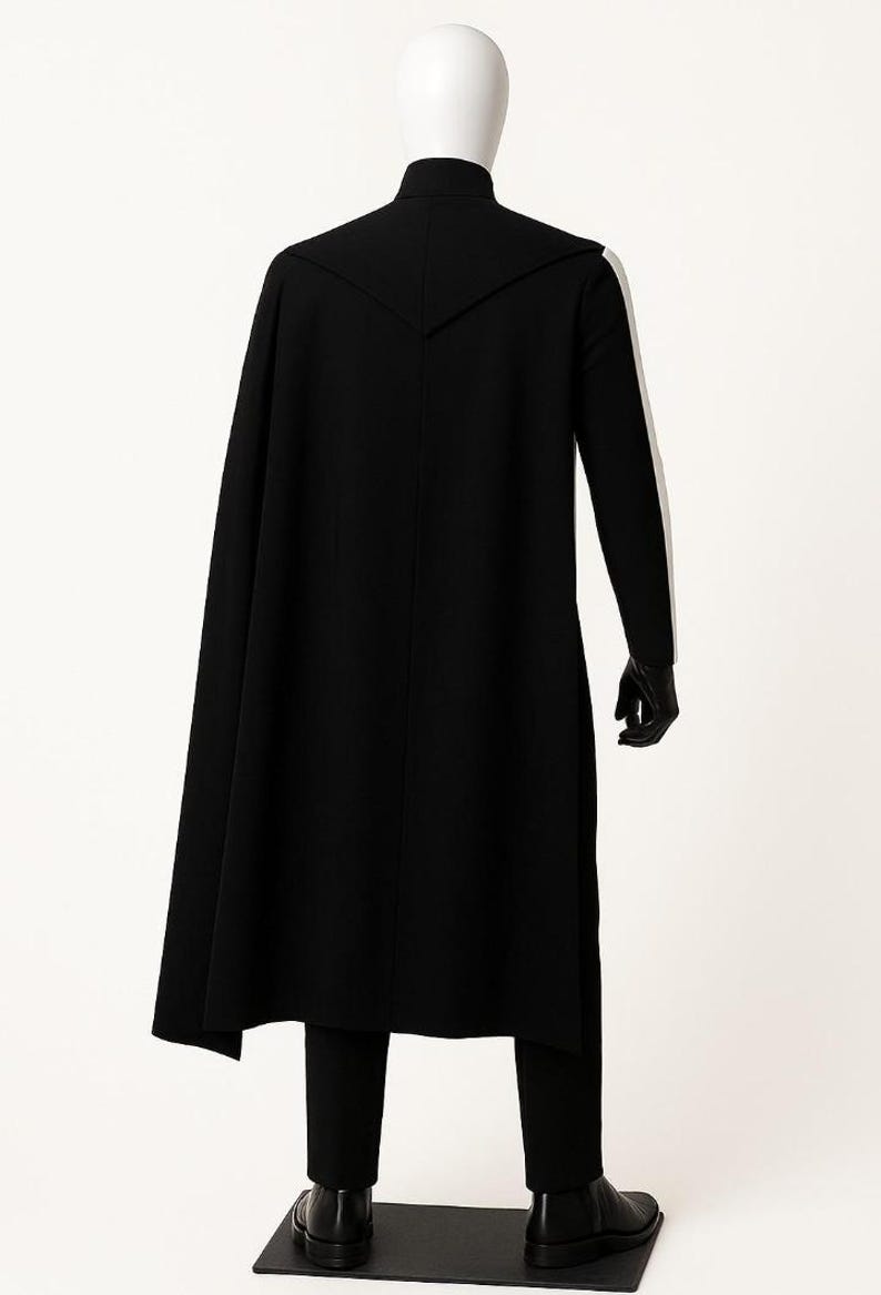May include: A black long-line coat with a high collar and a cape-like back panel, worn over matching black trousers. The ensemble includes black gloves and black boots, displayed on a mannequin against a plain white background.
