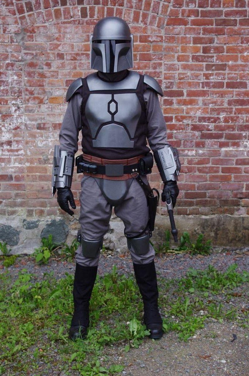 May include: A full-body costume resembling a Mandalorian warrior. The outfit includes a silver helmet, chest armor, and knee pads. The costume is gray and black, with a brown belt and black boots. A blaster pistol is visible.