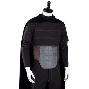 Pode incluir: Um conjunto de traje preto composto por uma capa longa, uma camisa de manga comprida com costuras verticais subtis e uma pe&ccedil;a de peito em camadas. O tronco inclui um painel abdominal retangular cinzento-escuro acolchoado sobre cal&ccedil;as pretas.