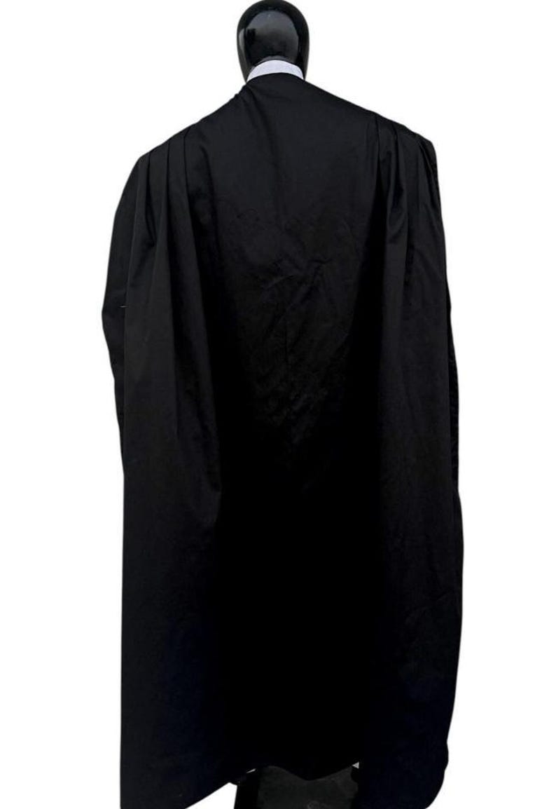 May include: A long, floor-length black cape featuring a high collar and a structured, pleated design. The garment is draped over a mannequin, showing a solid black fabric with a smooth texture and a clean, minimalist silhouette from the back view.