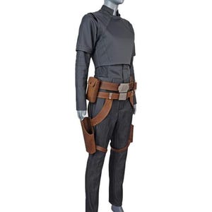 May include: A dark gray long-sleeved top and matching trousers worn with a brown leather utility belt featuring a holster and pouches. The ensemble includes a high-neck shirt with structured shoulders and a belt with metallic silver buckles.