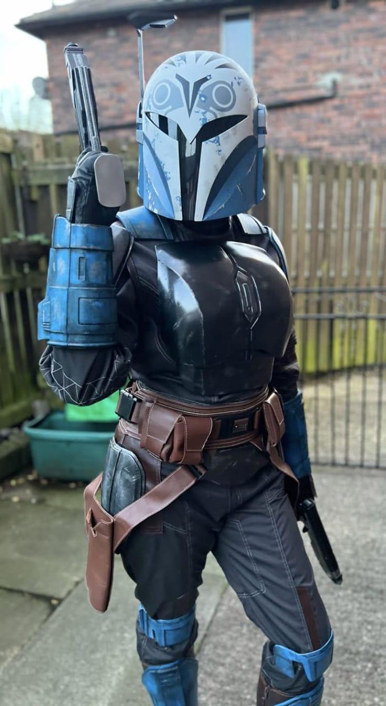 K&ouml;nnte beinhalten: Ein vollst&auml;ndiger R&uuml;stungsanzug mit einem blau-wei&szlig;en Helm mit stilisiertem Eulenmotiv, einer schwarzen Brustplatte, blauen Handschuhen und einem braunen G&uuml;rtel mit Holster. Das Set umfasst eine dunkle Hose und dazu passende blaue Kniepanzerungen.
