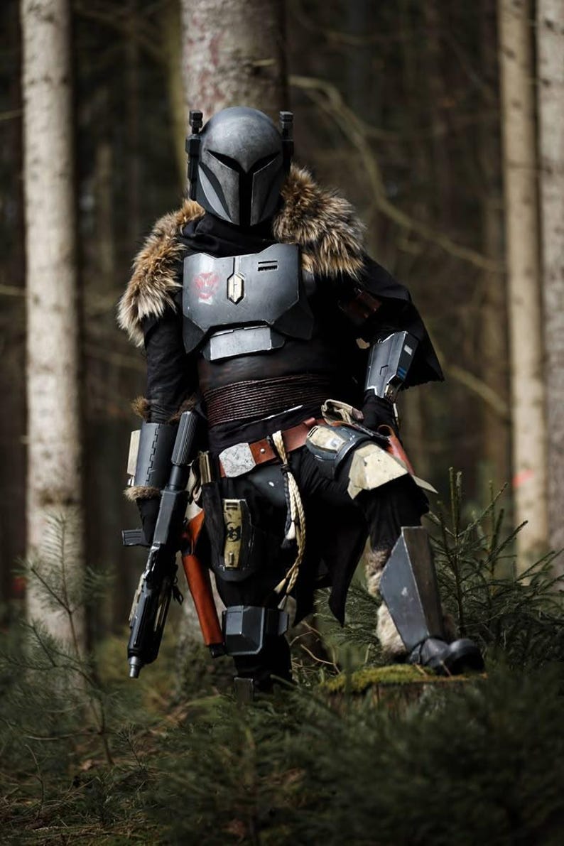 May include: A full-body suit of armor featuring a dark gray helmet with a T-shaped visor, a chest plate with a red emblem, brown fur shoulder accents, black fabric layers, metallic arm guards, a utility belt, and leg plating, holding a black blaster.