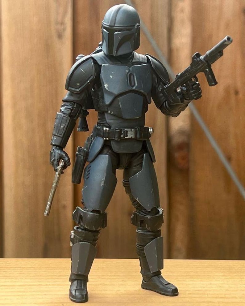 May include: A dark gray and black action figure wearing a full suit of armored plating, including a helmet with a T-shaped visor, chest plate, shoulder pauldrons, and leg guards. The figure holds a small blaster pistol in each hand.
