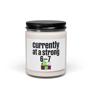 May include: A white candle jar with a black lid. The label reads "currently at a strong 6-7" with a green cartoon figure at a desk. The candle is a humorous gift.