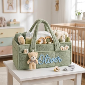 Puede incluir: Un organizador de pañales de tela texturizada en color verde salvia con dos asas, que presenta el nombre Oliver bordado en azul y un pequeño oso de peluche sujeto al frente. El organizador contiene artículos de bebé, incluyendo un biberón y juguetes.