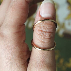 May include: Close-up of two thin, gold rings worn on a thumb. The rings are simple bands, likely made of gold or a gold-colored metal. The skin of the thumb is visible, showing fine lines and texture.