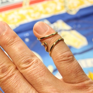 May include: A gold-colored finger ring with a spiral design. The ring wraps around the finger, with a band at the top and a curved, decorative element. The ring is worn on a finger.