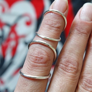 May include: A hand displaying four silver rings on one finger. The rings are delicate and plain, with a polished surface. The background is out of focus, featuring a red and white heart design.