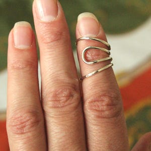 May include: A silver-colored finger ring with a unique, open design. The ring wraps around the finger with a series of curved and straight lines, creating an abstract, modern look. The ring is shown on a finger with a natural nail. Jewelry.
