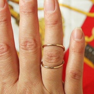 Può includere: Due sottili anelli color oro sono indossati su un dito. Gli anelli sono semplici fasce e il dito è posizionato su uno sfondo rosso e oro a motivi. Gli anelli sembrano essere di metallo.