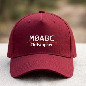 May include: A maroon baseball cap featuring the white text MØABC above a horizontal dashed orange line and the white name Christopher centered below. The cap has a structured crown, a curved brim, and embroidered eyelets on the panels.