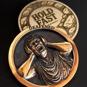 Puede incluir: Una moneda de color bronce con el relieve de una persona tapándose los oídos y gritando, con la palabra "DEAFENED" debajo. La moneda está enmarcada en color dorado. Una segunda moneda con las palabras "HOLD FAST" es visible en el fondo.