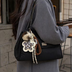 May include: Black shoulder bag with a rectangular shape and a flap closure. The bag features a decorative flower-shaped charm with a leopard print center and a fluffy pom-pom. The strap is black with a glossy finish.
