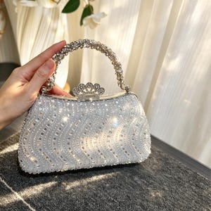 Può includere: Una borsa da sera argentata con un motivo ondulato e scintillante di strass. Il manico e la chiusura sono anch'essi decorati con cristalli. La borsa è tenuta in mano, indicando le sue dimensioni e l'idoneità come accessorio per occasioni speciali.