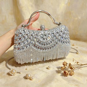 Puede incluir: Un bolso de mano plateado para la noche con asa y correa de cadena. El bolso está muy adornado con pedrería transparente y tiene una franja de cristales colgantes. Perlas y flores secas están esparcidas.