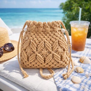 May include: A beige macrame bucket bag with a drawstring closure and a decorative floral pattern. The bag has a short handle and a long shoulder strap. The bag is sitting on a white surface with a straw hat, sunglasses, and a drink with a straw.