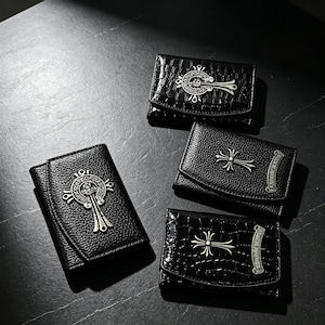 May include: Four black leather wallets with silver cross and decorative accents. Two wallets have a crocodile texture. The wallets are arranged on a dark surface, with a focus on the details of the design.