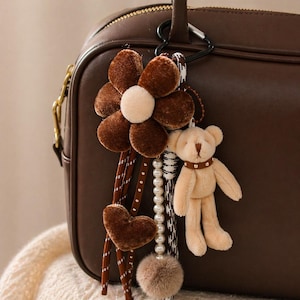 Pode incluir: Um chaveiro em forma de flor marrom com um centro creme, um coração marrom e um pequeno pingente de ursinho de pelúcia. O chaveiro também apresenta uma fileira de pérolas e um pompom fofo, preso a uma bolsa marrom.