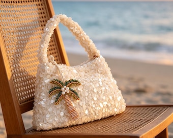 Puede incluir: Un bolso de mano color crema cubierto de lentejuelas, con un diseño de palmera con cuentas. El bolso tiene forma redondeada y un asa. Descansa sobre una silla de madera con asiento tejido, con el océano al fondo.