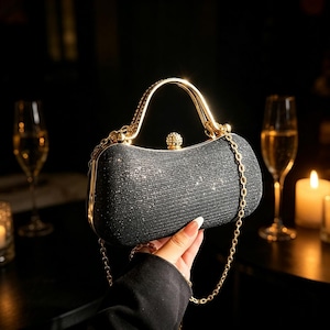 Könnte beinhalten: Eine schwarze, mit Glitzer bedeckte Clutch mit goldenem Griff und Kette. Die rechteckige Tasche hat eine abgerundete Oberseite und einen kleinen, dekorativen Schmuckstein am Verschluss. Die Clutch wird vor einem dunklen Hintergrund gehalten.