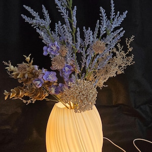 May include: A glowing, cream-colored ceramic vase with a spiral design, holding a bouquet of lavender and dried flowers. The vase sits on a wooden base, with a white cord extending from the back. The background is black.