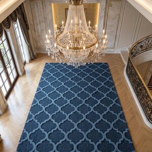 May include: A large rectangular area rug featuring a deep navy blue background with a raised, light blue lattice pattern in a quatrefoil design. The rug is positioned on a light-coloured wooden floor beneath a large, multi-tiered crystal chandelier.