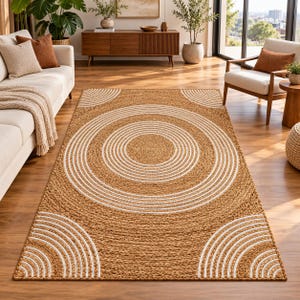 Op de afbeelding: Een rechthoekig vloerkleed gemaakt van gevlochten natuurlijke vezels in een beige kleur. Het kleed heeft een groot centraal patroon van concentrische witte cirkels en vier kleinere witte halve cirkelpatronen op de vier hoeken.