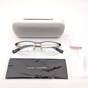 May include: Marc Jacobs semi-rimless spectacles with a brown and gold frame, featuring stainless steel construction. Includes a white hard case, a black microfibre cleaning cloth with white text, and a small plastic bag labelled Parts Package.