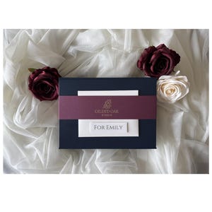 May include: A rectangular navy blue gift box featuring a burgundy paper band with the gold logo Gilded Oak Studio and a white rectangular label with the text FOR EMILY in black capital letters, centered on the box.