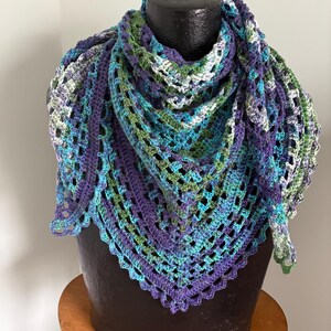 May include: A handmade crochet shawl in shades of blue, green, and purple. The triangular shawl is draped over a black mannequin head. The open weave design features a scalloped edge. The shawl is displayed on a wooden stool.