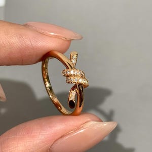 May include: A gold-toned ring featuring a knot-shaped design at the center. The knot is embellished with small, clear, sparkling stones. The band is smooth and polished, with the knot ends extending slightly beyond the main band structure.