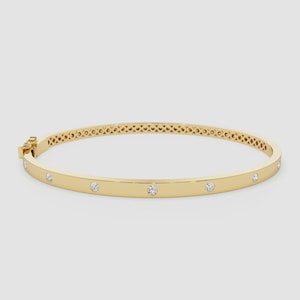 May include: A gold-coloured bangle bracelet featuring evenly spaced, small, clear gemstones. The bracelet has a clasp closure and a decorative openwork design on the top half. The bracelet's diameter is approximately 6.5 cm.