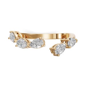 Op de afbeelding: Een open gouden ring met vijf witte, peervormig geslepen stenen langs de band. Het ontwerp heeft een opening tussen de stenen, met drie peervormige stenen aan de ene kant en twee aan de andere kant, gezet in een gepolijste gouden metalen band.