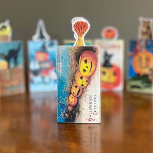 Può includere: Cinque cartoline vintage di Halloween con immagini diverse. Le cartoline sono impilate l'una sull'altra. La cartolina in cima presenta una fila di zucche con il testo "Halloween Greeting".