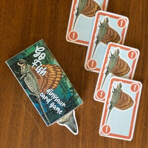 Puede incluir: Una caja de cartas de Go Fish de dinosaurios con una ilustración colorida de un dinosaurio en la parte delantera. La caja está sobre una mesa de madera con cuatro cartas que muestran un dinosaurio con el número 1 en cada carta.