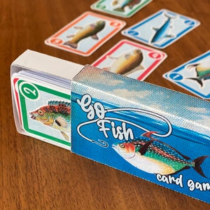 Puede incluir: Una caja azul con las palabras "Go Fish" y un anzuelo. La caja está abierta y muestra una baraja de cartas con ilustraciones de peces coloridos. Las cartas están numeradas.