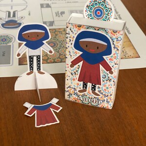 Puede incluir: Un conjunto de muñeca de papel con una niña que lleva un vestido rojo y un pañuelo azul. La muñeca está de pie sobre una base blanca con un fondo de patrones coloridos. El conjunto incluye un vestido separado y una pieza de cabeza con un diseño de patrones coloridos. El texto "Aaela" está impreso en la base.