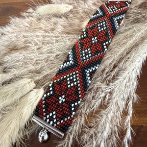 May include: A beaded bracelet with a geometric pattern in red, black, white, and gray. The bracelet has a silver-tone clasp and a small, dangling charm. The bracelet is approximately 1.5 inches wide.